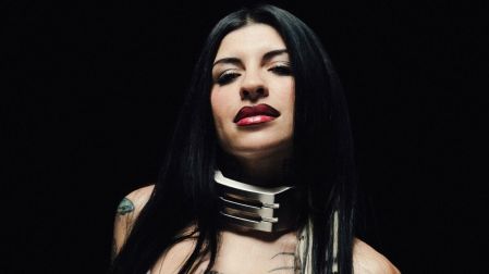 Cazzu con cabello negro largo, labios rojos, collar metálico plateado y tatuajes visibles sobre un fondo negro.