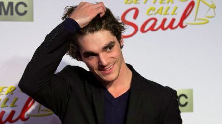 RJ Mitte con saco negro y camiseta azul oscuro, sonriendo y tocándose el cabello ante un fondo de Better Call Saul.