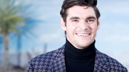RJ Mitte, actor de Breaking Bad, sonríe con saco azul estampado y suéter negro de cuello alto frente a un fondo azul.