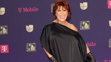 Alejandra Guzmán con cabello rojizo y vestido negro de un hombro, sonriendo ante un fondo morado de premios musicales.
