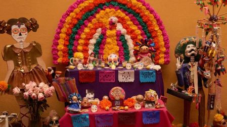 Ofrenda de Día de Muertos de dos niveles con flores multicolores, papel picado, calaveras, fotografías y figuras de catrinas.