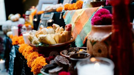 Ofrenda de Día de Muertos con tamales en una cazuela de barro, veladoras, flores de cempasúchil y otros alimentos tradicionales.