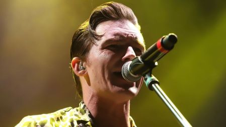 Drake Bell canta frente a un micrófono durante un concierto, con camisa estampada e iluminación amarilla en el escenario.