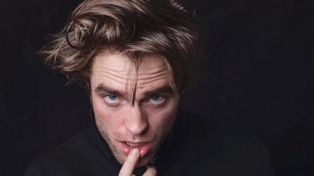 Robert Pattinson con cabello despeinado, ropa negra y un dedo sobre el labio inferior, frente a un fondo oscuro.