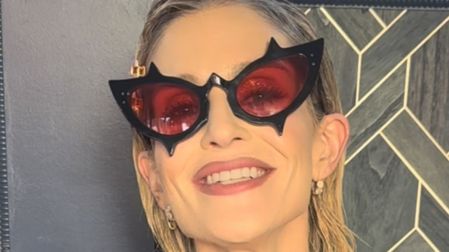 Kelly Osbourne sonríe con lentes negros de forma puntiaguda y cristales rojos, cabello rubio corto y aretes dorados.