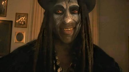 Personaje de American Horror Story 13 con rostro oscuro, ojos rojizos, cabello largo y sombrero negro en una escena de terror.