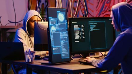 Dos personas con sudaderas y capuchas frente a varias computadoras con pantallas de código, relacionadas con hackeo en modo incógnito.