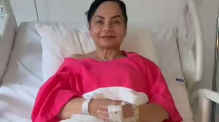 Bettina Salazar, Olga Sana, recostada en una cama de hospital con bata rosa, pulseras médicas y las manos sobre una sábana blanca.