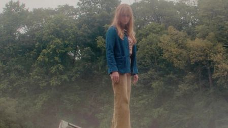 Pamela Voorhees en Crystal Lake, con cabello rubio, chaqueta azul y pantalón beige, de pie sobre un muelle frente a un bosque.