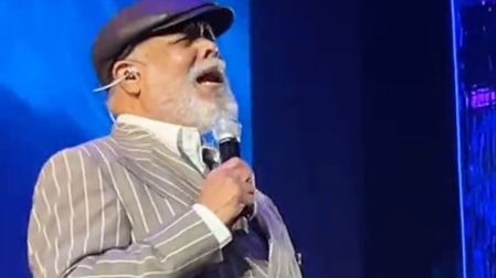 Francisco Céspedes canta con micrófono, traje gris de rayas y gorra negra sobre un escenario iluminado de azul.