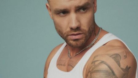 Liam Payne con camiseta blanca sin mangas, cadena plateada, barba corta y tatuajes en el hombro, frente a un fondo azul claro.