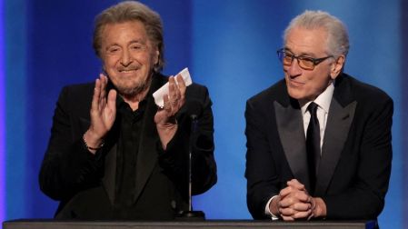 Al Pacino aplaude junto a Robert De Niro, quien viste traje negro y lentes, durante una presentación sobre un escenario azul.