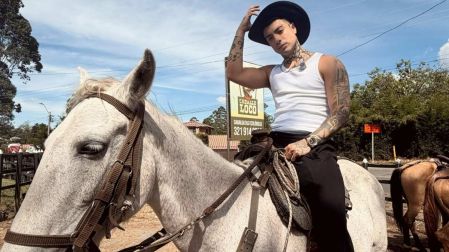 Naim Darrechi posa a caballo sobre un ejemplar blanco, con sombrero negro, camiseta sin mangas y tatuajes visibles.
