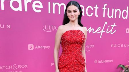 Selena Gomez posa con un vestido rojo de pedrería y cabello negro suelto ante un fondo rosa en un evento benéfico.