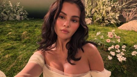 Selena Gomez se toma una selfie con vestido blanco y cabello oscuro ondulado frente a un jardín con flores.