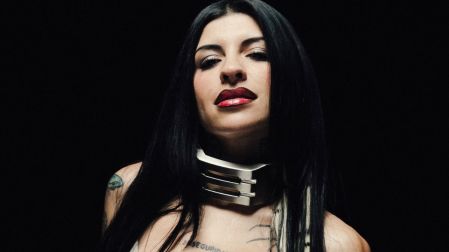 Cazzu posa con cabello negro largo, labios rojos, collar metálico y tatuajes visibles sobre un fondo negro.
