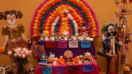 Ofrenda de Día de Muertos con flores multicolores, papel picado, calaveras decorativas, muñecas de catrinas y fotografías.