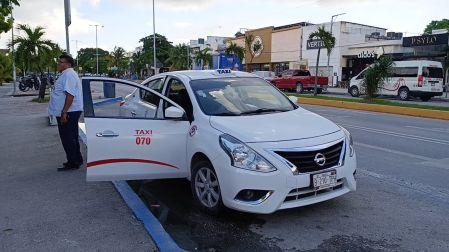Conductor de taxi blanco número 070 parado junto a su vehículo con la puerta abierta esperando pasajeros en Tulum.