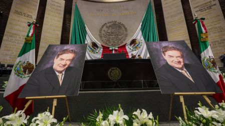 Retrato del periodista Francisco Garfias Antolín junto a la bandera de México y flores blancas en San Lázaro.