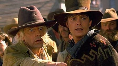 Doc Brown señala hacia el frente junto a Marty McFly, ambos vestidos como vaqueros en una escena de Volver al futuro III.