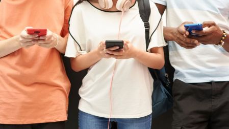 Tres jóvenes utilizan sus celulares de colores rojo, negro y azul; una lleva audífonos alrededor del cuello.