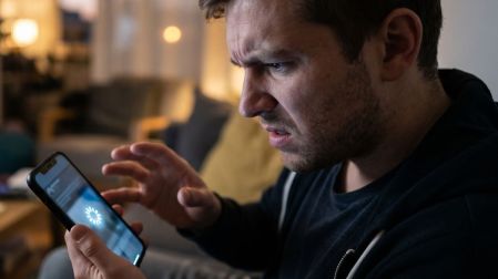 Hombre observa con frustración su celular congelado, cuya pantalla muestra un símbolo de carga sin avanzar.