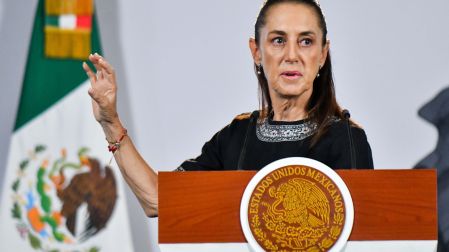 La presidenta Claudia Sheinbaum hablando desde el atril presidencial con el escudo de los Estados Unidos Mexicanos, teniendo la bandera de México de fondo.