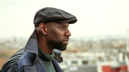 Omar Sy como Assane Diop en Lupin 3, con boina negra, abrigo oscuro y suéter verde, mirando de perfil.