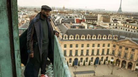 Omar Sy como Assane Diop en Lupin, con boina y abrigo oscuro sobre un balcón de París, con la Torre Eiffel al fondo.