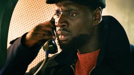 Omar Sy como Assane Diop en Lupin 3, con boina negra y suéter rojo, hablando por teléfono en una cabina