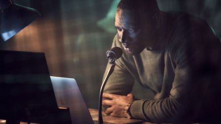 Omar Sy como Assane Diop en Lupin 3, inclinado frente a una computadora portátil y un micrófono sobre un escritorio.