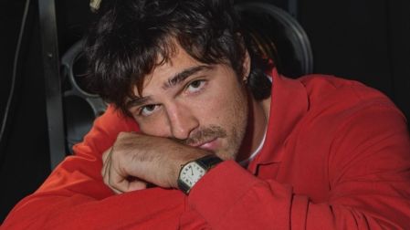 Jacob Elordi con cabello oscuro y camisa roja, apoyando el rostro sobre sus brazos y mirando a la cámara.