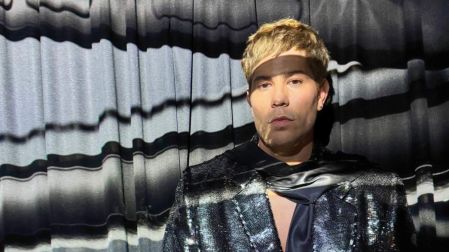 Christian Chávez luce cabello rubio y saco negro brillante frente a un fondo de cortinas negras y grises.