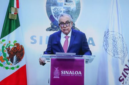 Rubén Rocha Moya, Gobernador de Sinaloa, en conferencia de prensa. Viste traje azul, corbata fucsia y gafas.