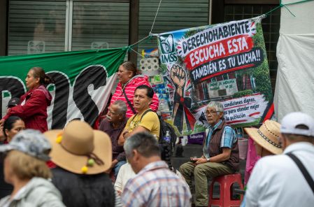 arias personas sentadas y reunidas en un plantón debajo de una lona que dice "Derechohabiente escucha esta también es tu lucha!!" junto al logotipo del IMSS.