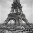 Fotografía histórica de la Torre Eiffel en construcción en el Campo de Marte, París, con obreros, andamios y los cuatro pilares de hierro visibles mientras la estructura se eleva antes de su inauguración en 1889.