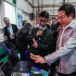 Estudiante explica proyecto tecnológico con laptop a autoridad educativa durante actividad de educación dual en espacio de exhibición con docentes.