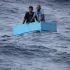 Dos hombres aferrados a una hielera rectangular de color azul claro, flotando a la deriva en el océano agitado.