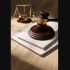 La imagen muestra un mazo de juez de madera con un mango dorado descansando sobre un conjunto de papeles o documentos apilados. Al fondo, parcialmente desenfocado, se ve una balanza de la justicia, un símbolo clásico que representa el equilibrio y la equidad en el sistema legal. Estos elementos están sobre una superficie de madera y la iluminación de la escena sugiere un entorno formal, posiblemente un despacho o sala de audiencias.