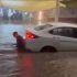 Sedán blanco detenido en una calle con agua a la altura de las ventanas traseras durante una fuerte tormenta en Ciudad de México. Un hombre camina junto al auto, sumergido hasta la cintura, en medio de una corriente que dificulta el paso y amenaza con daños severos al motor y sistemas eléctricos.
