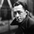 Retrato en blanco y negro de Albert Camus mirando a cámara, con abrigo oscuro y un cigarrillo en la boca, en un entorno urbano desenfocado que refuerza su imagen de escritor reflexivo y existencialista.