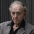 Retrato de Frederick Wiseman, documentalista estadounidense, con saco gris y camisa oscura, frente a un fondo textil. La imagen acompaña una nota sobre su fallecimiento a los 96 años y su legado en el cine documental.