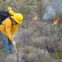 Brigadista con casco y uniforme amarillo trabaja con herramienta manual para contener un incendio forestal en zona de matorral en Múzquiz, Coahuila, mientras al fondo se observan llamas activas entre la vegetación seca.