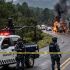 Una patrulla de la Guardia Nacional y agentes de la policía establecen un cordón de seguridad con cinta amarilla en una carretera de Michoacán. En el fondo, varios camiones de carga están incendiándose, emitiendo gruesas columnas de humo negro en un entorno boscoso y montañoso. Imagen representativa de narcobloqueos y violencia en la región.