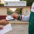 Funcionario de la SEP entregando la tarjeta del Banco del Bienestar para la Beca Rita Cetina a una madre de familia afuera de una escuela primaria.
