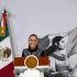 La presidenta Claudia Sheinbaum sonriendo en el podio durante su conferencia de prensa matutina en Palacio Nacional, con la bandera de México y la ilustración de una mujer indígena de fondo.