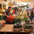 ista de un concurrido mercado al aire libre de México con letreros de precios. Una mujer mayor revisa un aguacate en primer plano con un cartel que dice 'Aguacate $75.00/Kg'. Al fondo, un vendedor sonríe frente a guacales de jitomates rojos ('Jitomate $15.00/Kg') y chiles serranos ('Chile Serrano $20.00/Kg'), ilustrando el costo real de los productos básicos reportados en el informe de inflación del Inegi