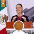 Una fotografía de primer plano de Claudia Sheinbaum, Presidenta de México, sonriendo ampliamente detrás del podio oficial del Gobierno de México con el Sello Nacional visible, vistiendo un traje oscuro con bordados de colores. La bandera nacional y un diseño artístico de fondo son visibles