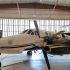 Vista exterior del avión Beechcraft blanco con franjas negras del Mayo Zambada exhibido en un hangar, con gruesas cadenas en la hélice derecha.