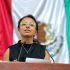 Diputada local con lentes amarillos habla desde la tribuna legislativa, frente a una bandera de México durante una sesión pública.